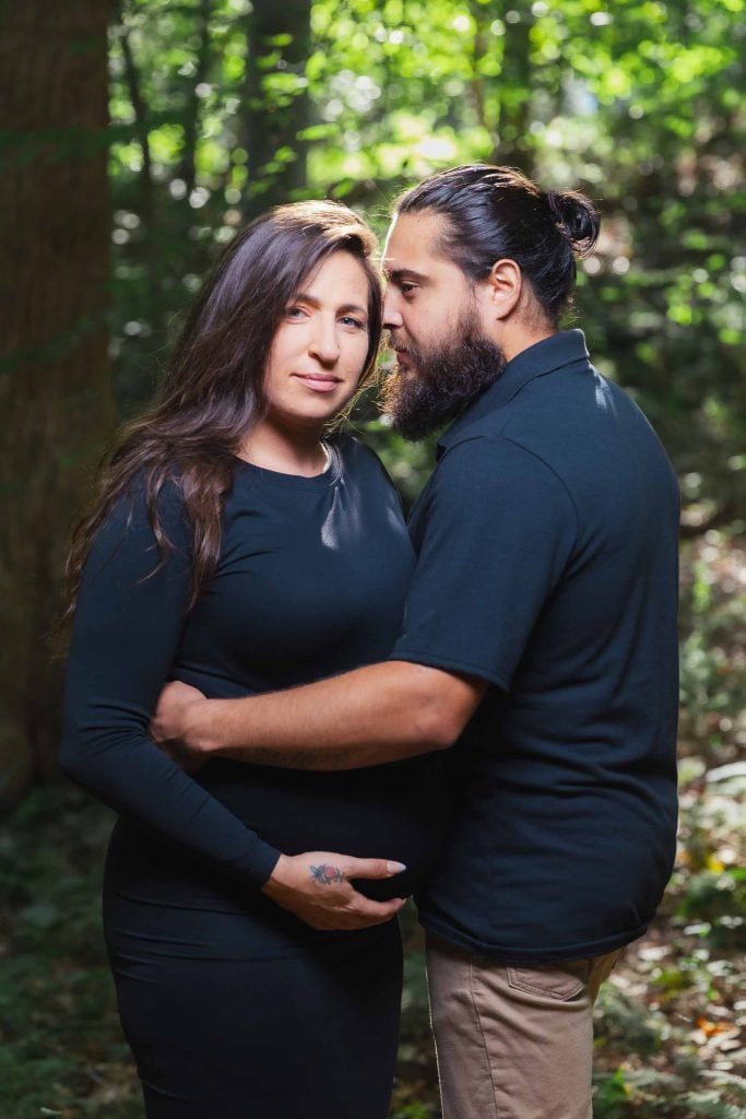 A man and a pregnant woman stand close together in a forest, sharing an intimate moment as he kisses her forehead—their hands resting on her belly in this tender outdoor maternity portrait.