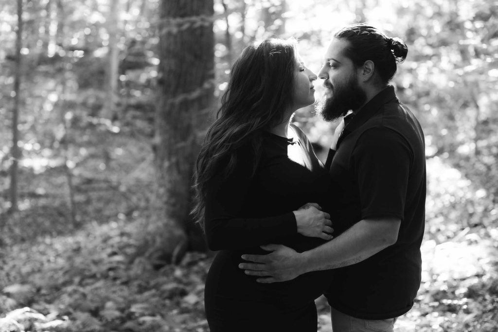 A man and a pregnant woman stand close together in a wooded area, gazing at each other with hands on her belly in this tender outdoor maternity portrait.