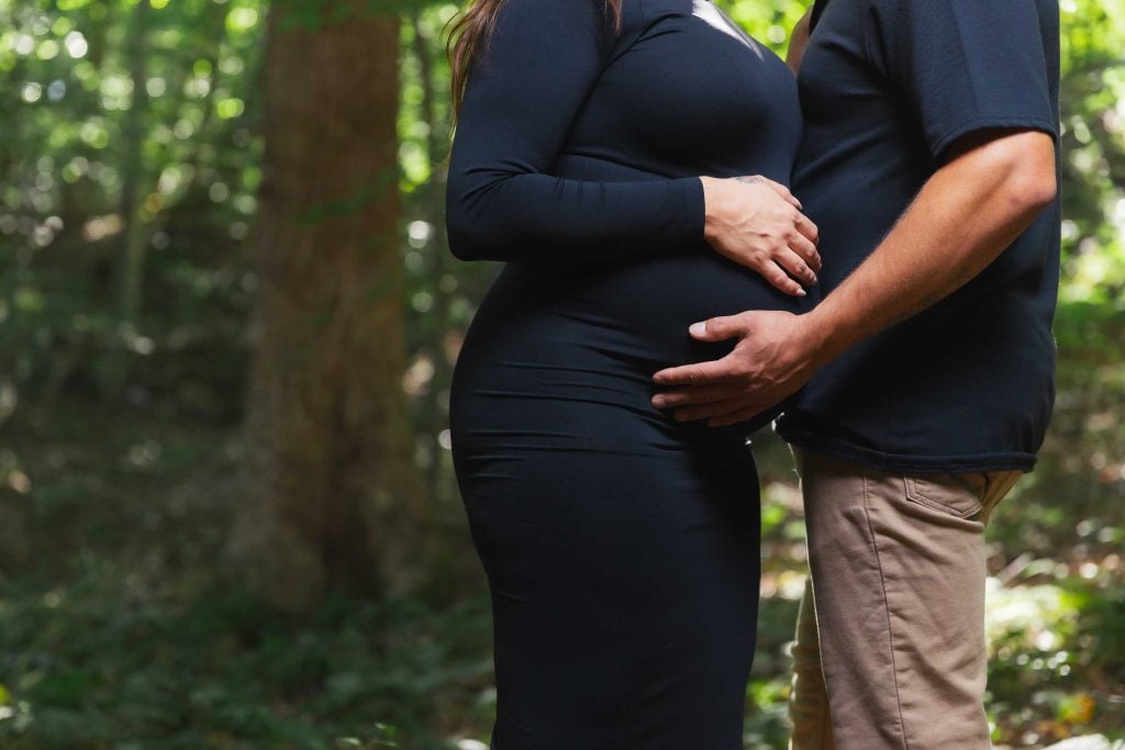 Two people stand in a wooded area, their hands gently resting on a pregnant belly. This outdoor maternity portrait captures only their torsos, emphasizing the joy and anticipation of new life.