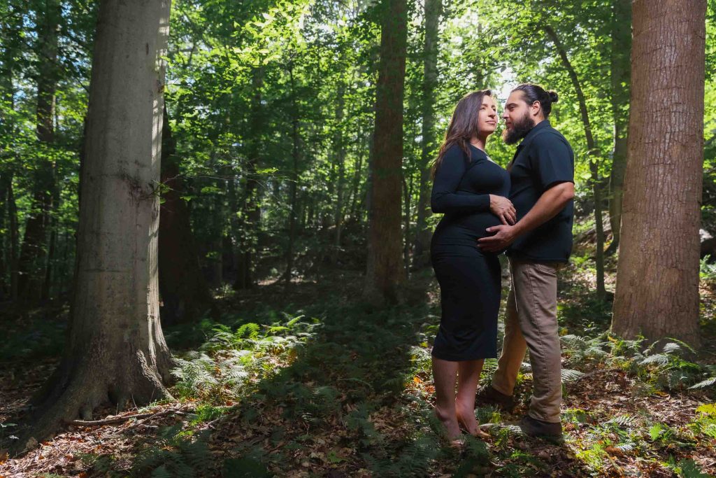 A couple stands together in a sunlit forest, the man gently placing his hand on the woman's pregnant belly—an intimate outdoor maternity portrait bathed in golden light.