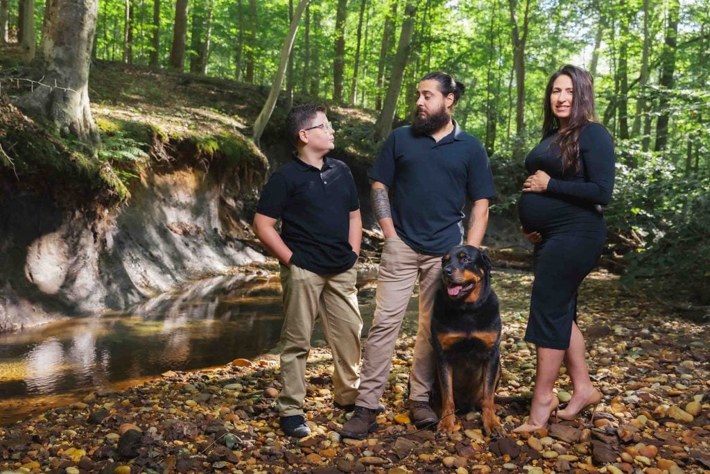 A family of three and a dog stand on a rocky creek bed in a forest; the woman appears pregnant, creating a heartfelt outdoor maternity portrait.