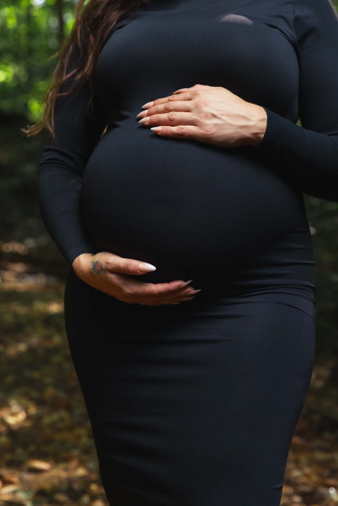 A person in a black long-sleeve dress holds their pregnant belly with both hands, captured in a serene outdoor maternity portrait.