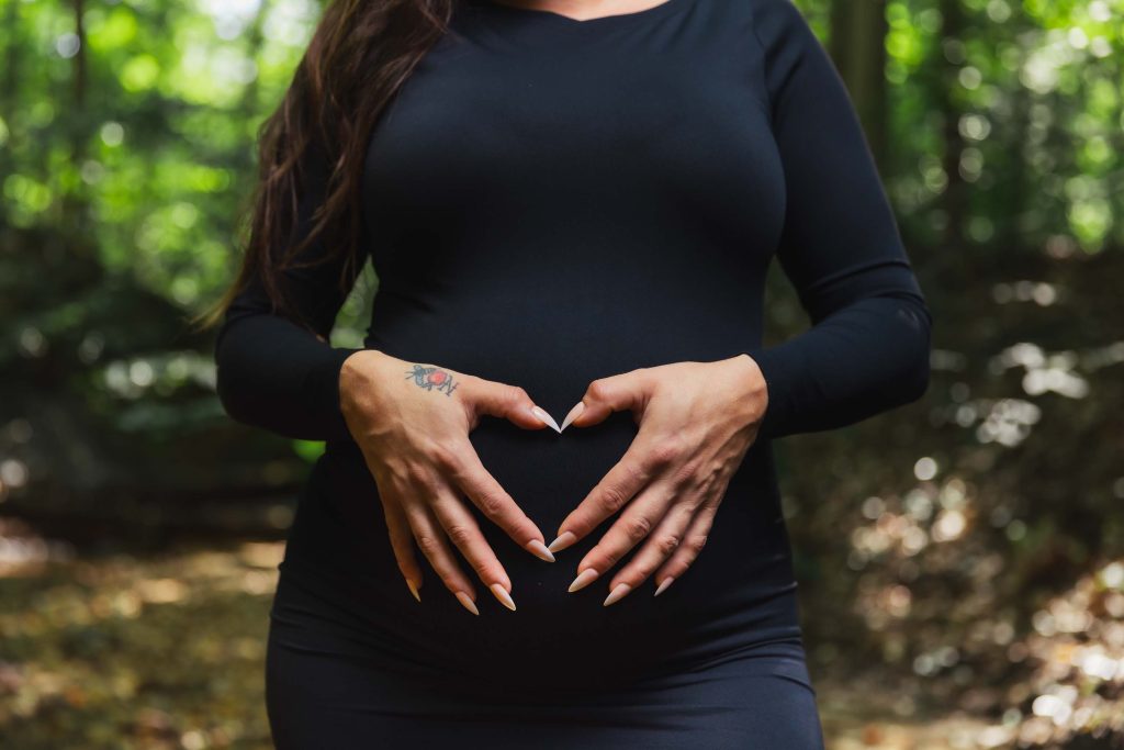 A person in a black dress stands in a forest, forming a heart shape with their hands over a pregnant belly—capturing the beauty of outdoor maternity portraits.