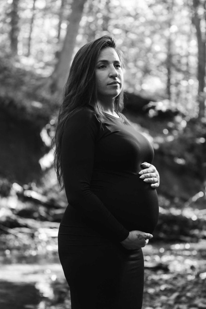 A maternity portrait of a pregnant woman in a long-sleeved dress standing outdoors in a wooded area, holding her belly and looking at the camera.