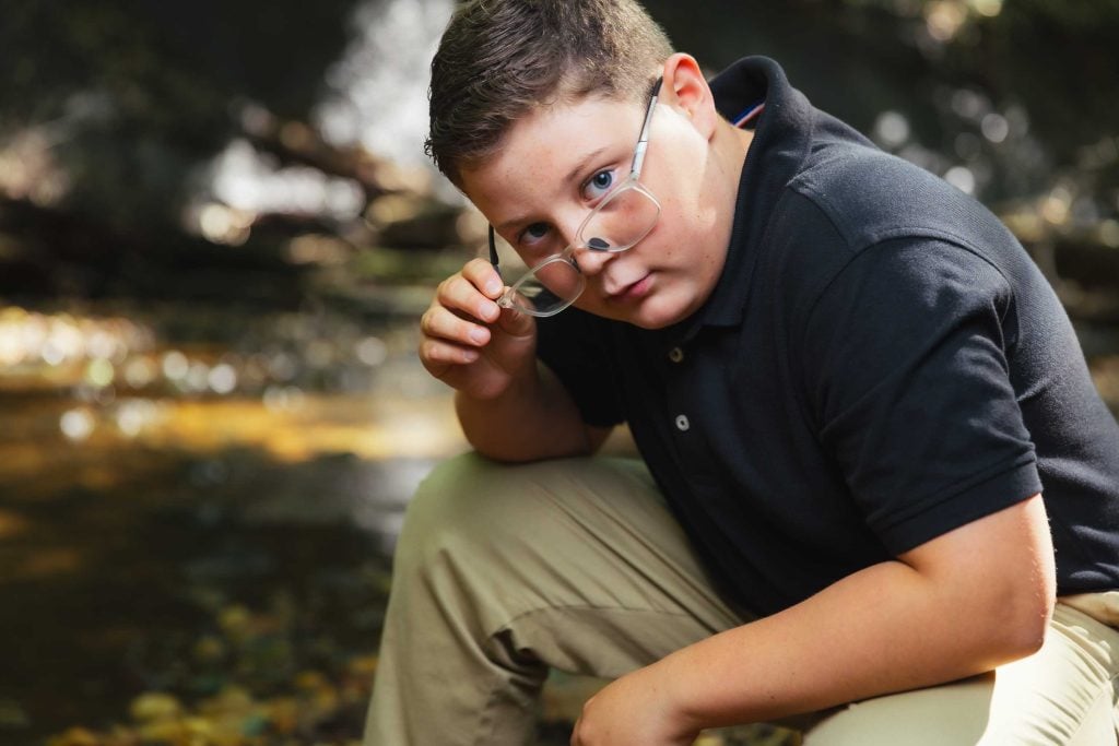A boy in a black polo shirt and khaki pants holds his glasses while looking at the camera, crouched outdoors near a stream—an ideal setting for relaxed outdoor portraits.