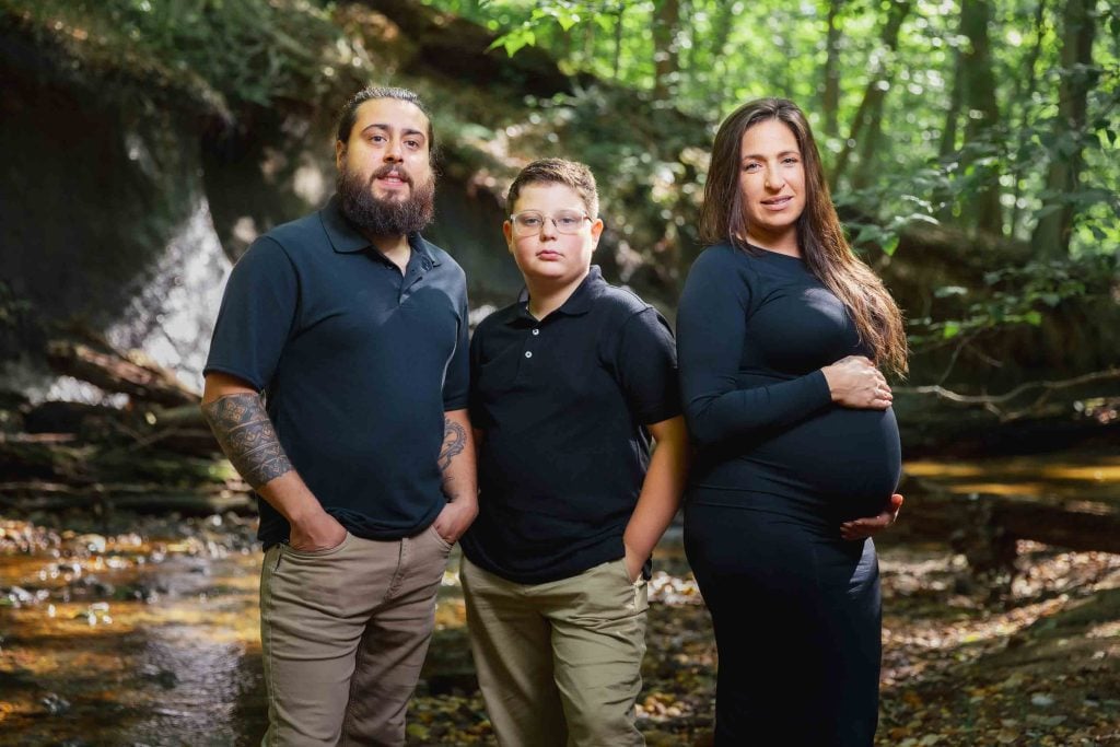 Three people, including a pregnant woman, a man, and a boy, stand side by side in a wooded area near a stream, dressed in dark tops and tan or black pants, capturing a serene outdoor maternity portrait.
