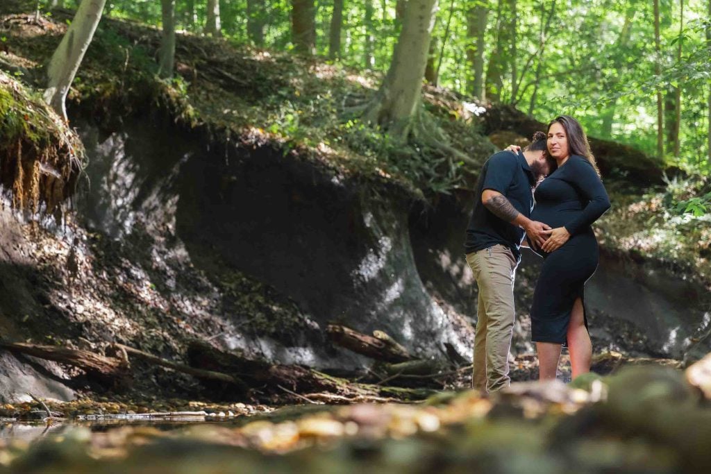 An outdoor maternity portrait of a man and a pregnant woman standing together in a sunlit forest, with the man gently touching her belly as they are surrounded by trees and dappled light.
