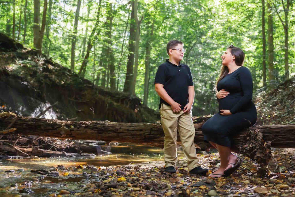 A man and a pregnant woman are talking and smiling by a fallen tree in a forest, enjoying an outdoor maternity moment with rocks and a stream in the background—capturing the essence of natural portraits.