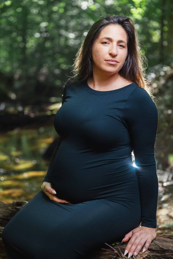 A pregnant woman in a black dress sits on a log in a sunlit forest near a stream, resting one hand on her belly and looking at the camera—an enchanting outdoor maternity portrait.