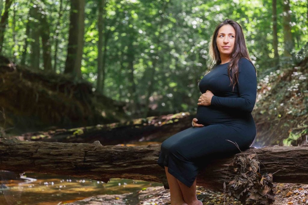 A pregnant woman in a long black dress sits on a fallen log in the forest, holding her belly and gazing into the distance—an elegant outdoor maternity portrait.