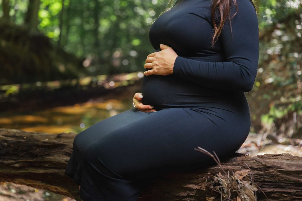 A maternity portrait shows a pregnant person in a long black dress, sitting on a fallen tree in the forest and gently holding their belly with both hands.