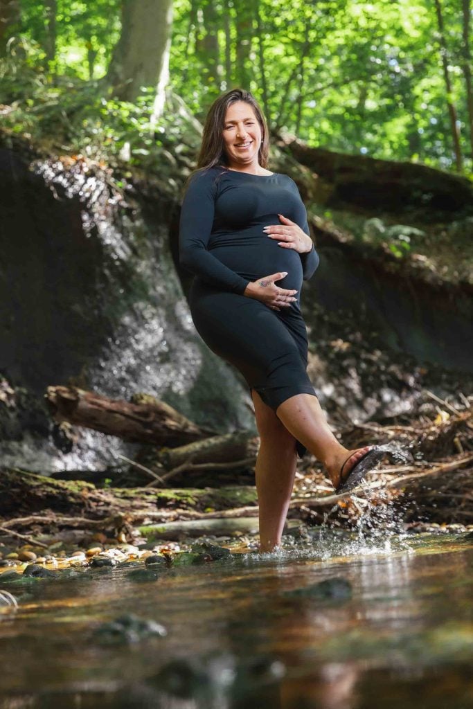This outdoor maternity portrait captures a pregnant woman in a black dress, smiling as she stands in a shallow stream, touching her belly and splashing water with one foot, surrounded by a serene forest.