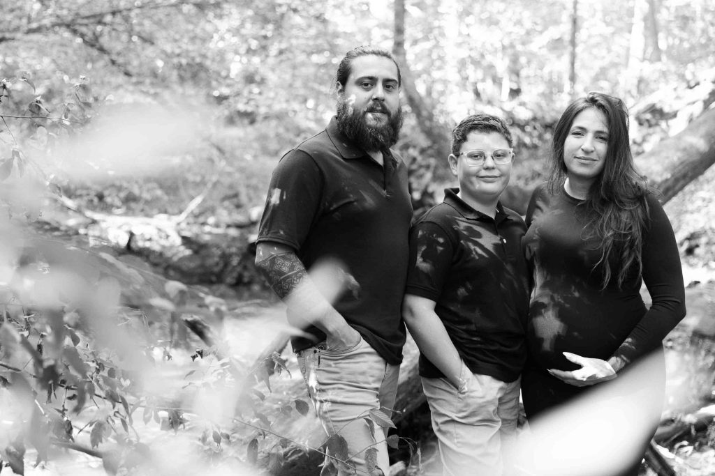 Three people stand side by side outdoors in a wooded area, posing for a black and white portrait. They are wearing dark shirts and light pants, creating a classic outdoor scene.